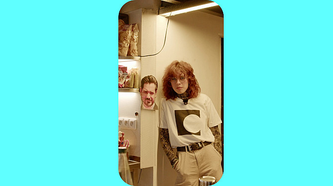 Žena se opírá ramenem o zeď, vedle ní visí na zdi mužská hlava vytištěná na papíře.