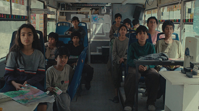 Skupina dětí sedí uvnitř upraveného autobusu sloužícího jako mobilní učebna, dívají se směrem dopředu. Některé mají na lavicích sešity a školní pomůcky, interiér je zaplněný knihami a vzdělávacími materiály.