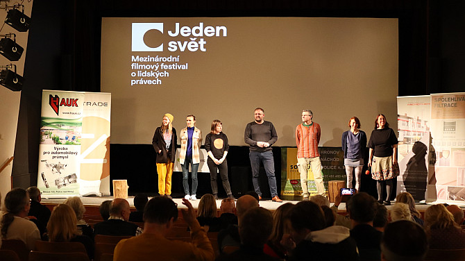 Na pódiu kina stojí v řadě osm lidí před velkým promítacím plátnem s logem festivalu Jeden svět a textem o mezinárodním filmovém festivalu o lidských právech. Po stranách jsou reklamní bannery. V popředí sedí diváci v hledišti a sledují probíhající diskusi.