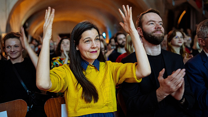 Jen titulky nestačí. Jeden svět slaví Mezinárodní týden neslyšících a ukazuje, že přístupnost není jen prázdné gesto Návštěvníci sedí v zaplněném sále a tleskají během kulturní akce. V popředí je osoba ve žlutém svetru se zdviženýma rukama, která tleská znakovou řečí.