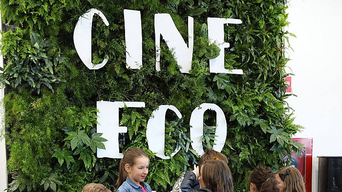 Udržitelnost na Jednom světě: Z plátna do praxe Skupina dětí stojí před zelenou stěnou pokrytou rostlinami s velkým nápisem ‚CINE ECO‘.