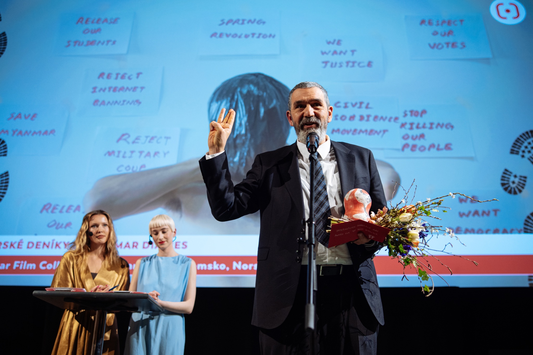 Na podiu stojí Igor Blaževič s cenou a kyticí v ruce. Za ním je na projekčním plátnu snímek z vítězného filmu.