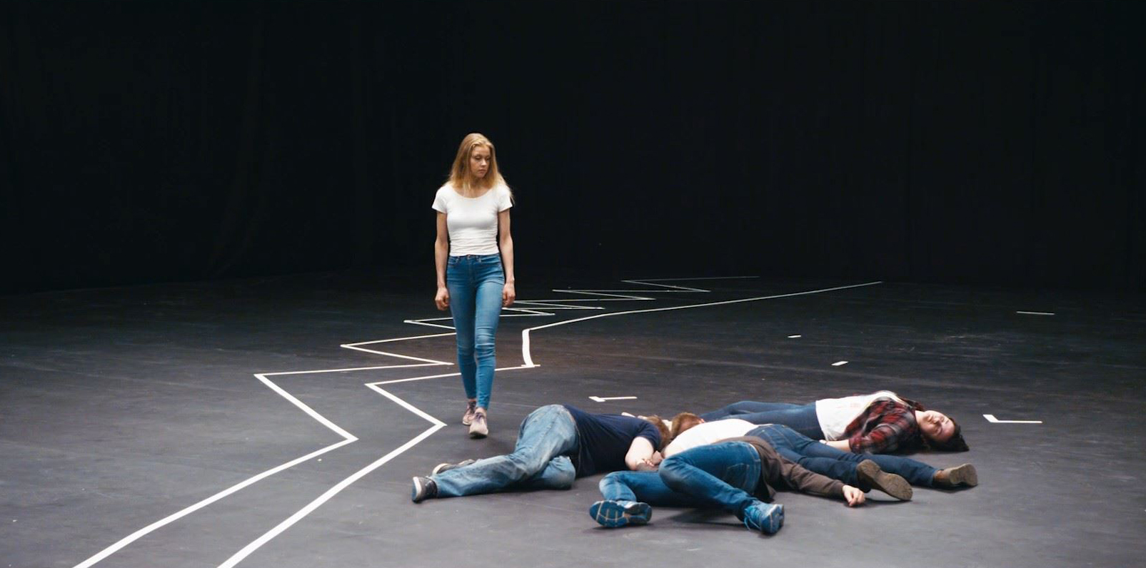 Na scéně bez pozadí stojí osoba a vedle ní leží na zemi čtyři osoby. Na podlaze jsou lepící páskou vyznačené různé linie.