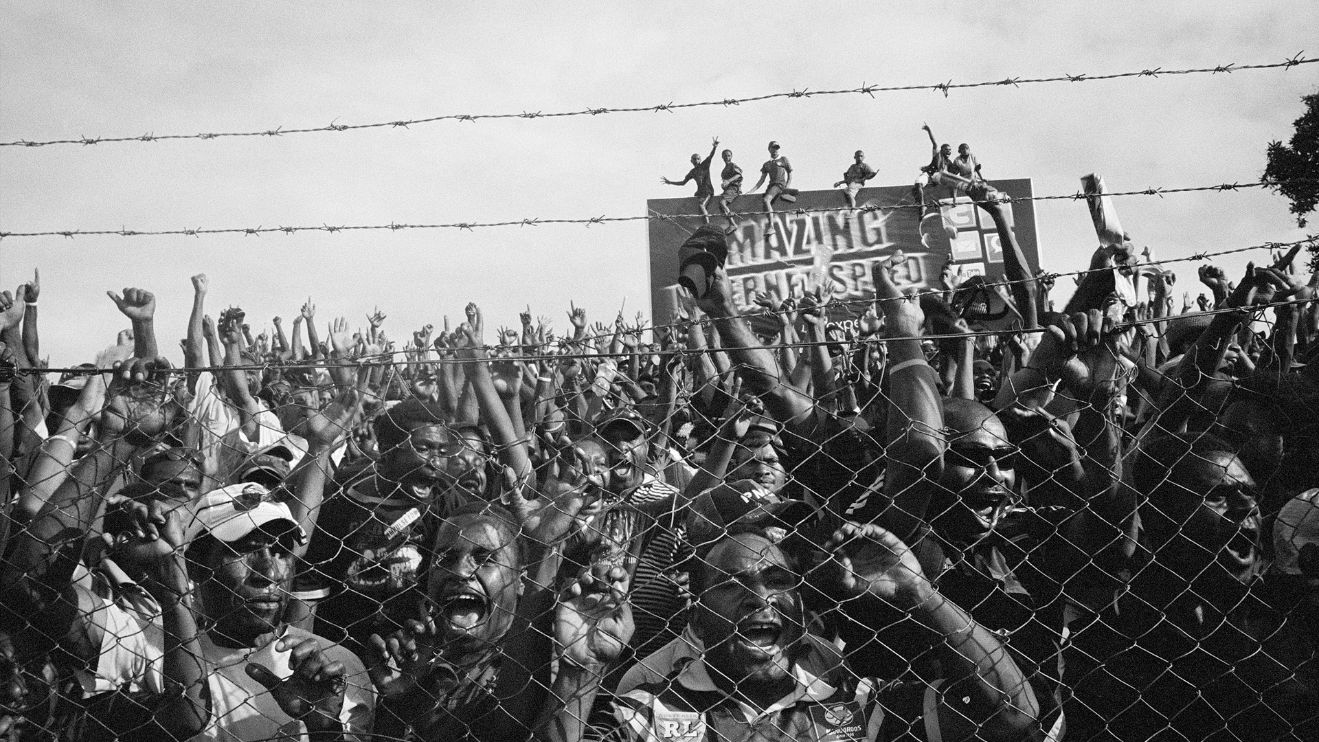 Za vysokým plotem, nad kterým vede ochranný drát z žiletek, stojí dav mužů s tmavou pletí. Křičí a zvedají ruce nad hlavu. / A crowd of dark-skinned men stand behind a high fence with razor wire running over it. They shout and raise their hands above their heads.