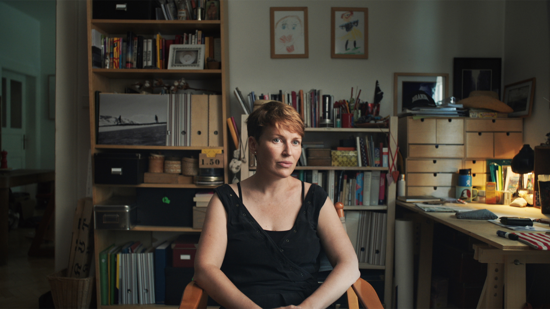 Investigativní novinářka Pavla Holcová sedí na židli uprostřed tmavé kanceláře. Vedle ní je stůl s papíry a za ní knihovna s šanony. / Investigative journalist Pavla Holcová sits on a chair in the middle of a dark office. Next to her is a desk with papers and behind her a bookcase with binders.