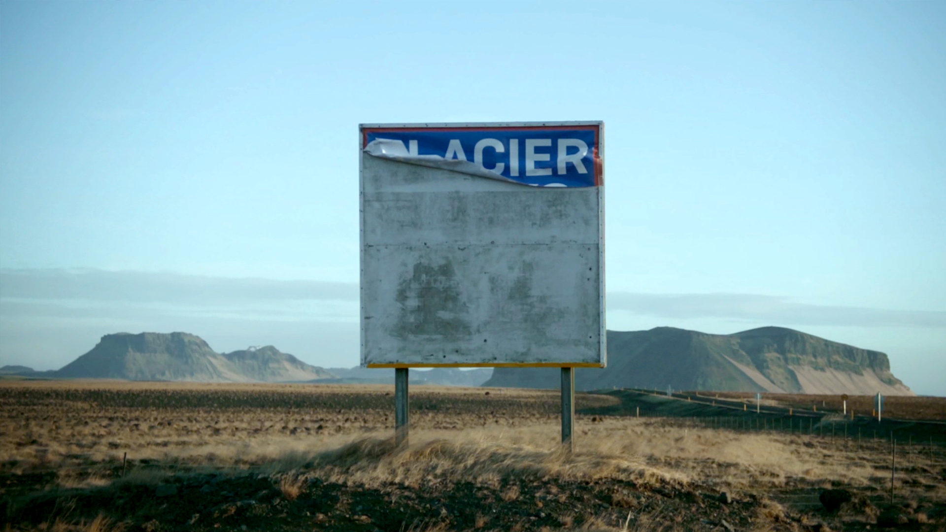 Uprostřed je starý ošuntělý billboard s nápisem ledovec. V pozadí jsou kopce a pláně islandské krajiny. / In the middle is a shabby old billboard with the word glacier on it. In the background are the hills and plains of the Icelandic landscape.