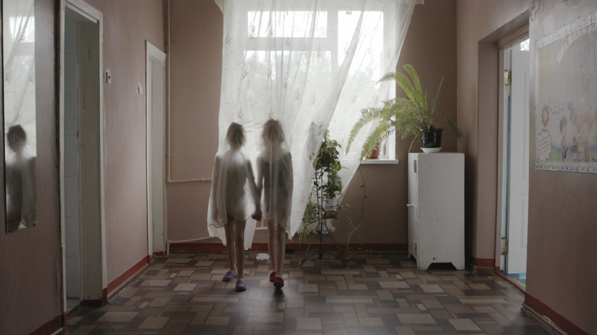 Dvě dívky se drží za ruce a stojí za záclonou na chodbě Donbaského sirotčince.