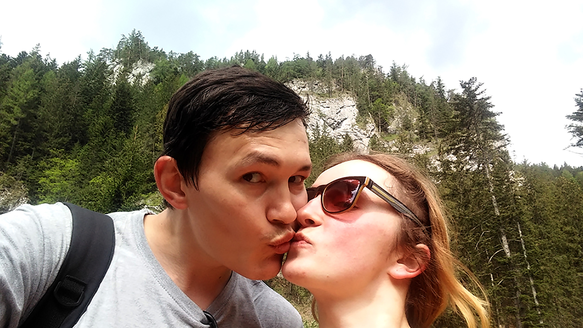 Na selfie se hlavní hrdinové líbají v přírodě s horami a stromy za zády.  / A selfie shows the protagonists sharing a kiss while out in nature with mountains and trees behind them.