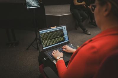 Právo na znakový jazyk je lidské právo Osoba zapisuje na notebooku simultánní přepis pro neslyšící účastníky debaty. V pozadí sedí lidé a na stojanu je vidět další monitor s promítaným přepisem.