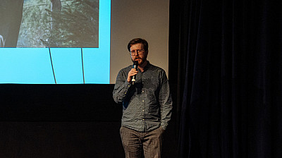 Muž stojící před filmovým plátnem mluví do mikrofonu.