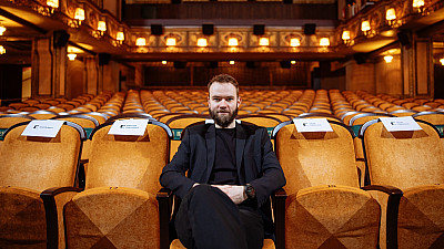 Muž sedí na sedadle uprostřed první řady prázdného kinosálu.