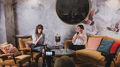 Dvě ženy sedí v pohodlném křesle a na pohovce v útulném interiéru a drží mikrofony během podcastového rozhovoru. Před nimi stojí malý stolek se sklenicemi vody a natáčení zaznamenávají kamery; v kulatém zrcadle na zdi je vidět odraz sedícího publika.