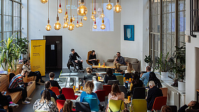 Panelová diskuse v moderním coworkingovém prostoru, kde tři řečníci sedí na pódiu před publikem; nad nimi visí dekorativní žárovky a za nimi je promítaná prezentace. Posluchači sedí na barevných židlích a sledují debatu.