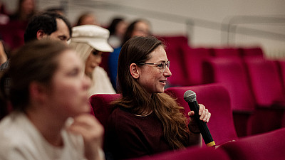 Žena sedící v červeném křesle drží mikrofon a pokládá otázku během diskuse. Má brýle a dlouhé hnědé vlasy, kolem ní sedí další účastníci, zatímco pozadí tvoří řady červených sedadel.