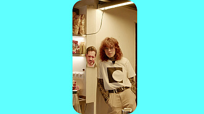 Žena se opírá ramenem o zeď, vedle ní visí na zdi mužská hlava vytištěná na papíře.