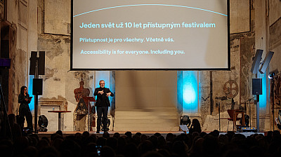 Na pódiu v historickém interiéru stojí muž, který tlumočí do znakového jazyka, vedle něj žena s poznámkami. Za nimi je velké projekční plátno s nápisem o festivalu Jeden svět a sdělením o přístupnosti pro všechny. Před pódiem sedí publikum sledující program.