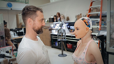 V dílně stojí člověk tváří v tvář humanoidnímu robotovi. Robot má odkrytou část hlavy s viditelnou elektronikou.