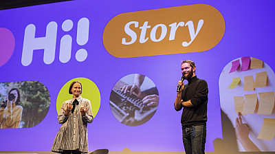 Na pódiu stojí dva přednášející před velkou projekční plochou s nápisem „Hi! Story“ a několika fotografiemi. 