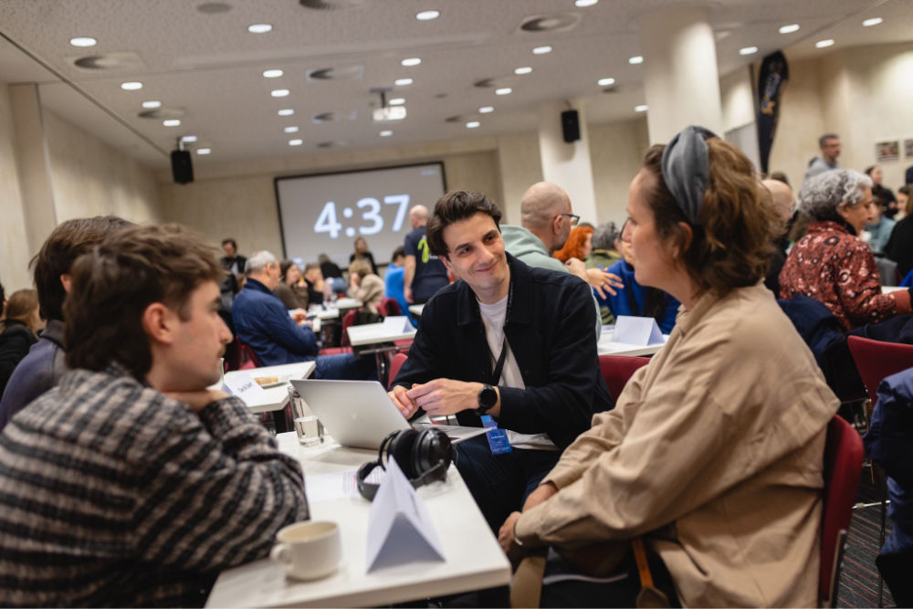 Skupina lidí sedí u stolu a diskutuje na konferenci, v pozadí je velká obrazovka s časem.