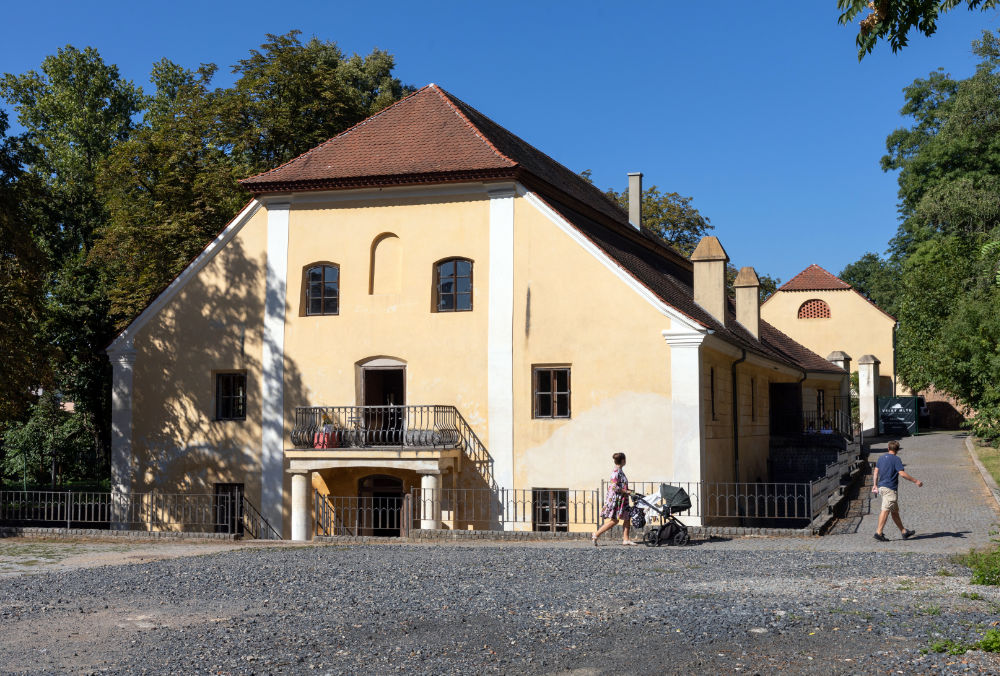 Žlutý historický dům s červenou střechou, před kterým procházejí lidé s kočárkem.