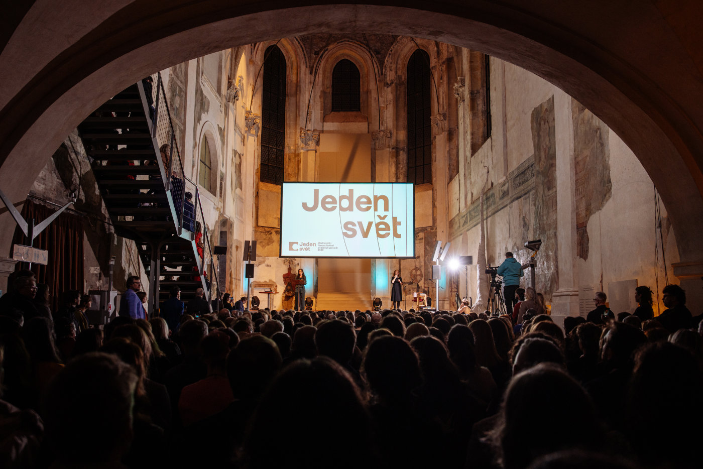 Promítací sál s historickými zdmi, plátno s nápisem „Jeden svět“ a diváci sedící v hledišti.