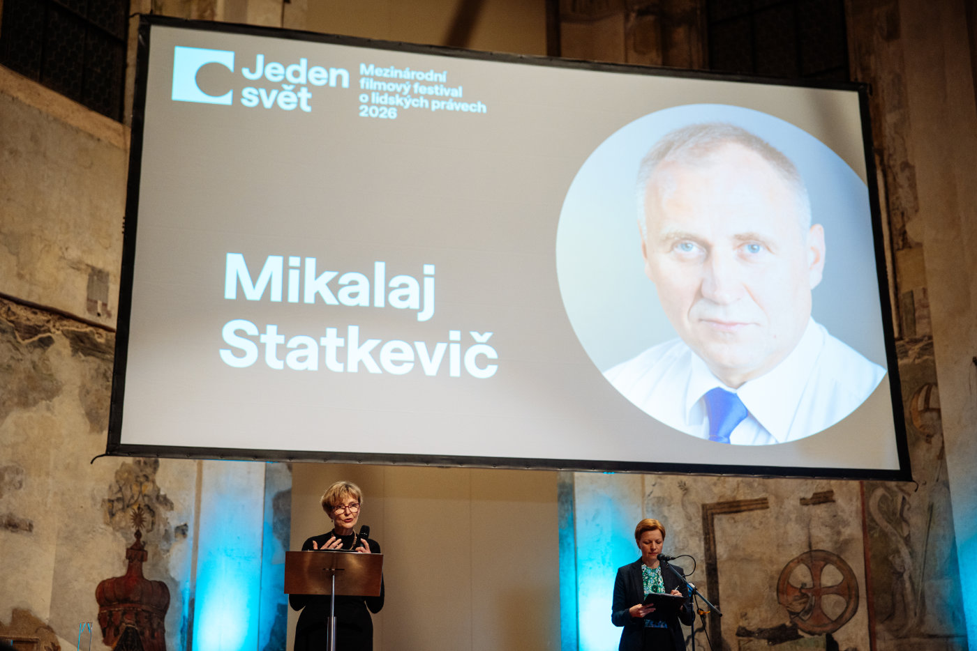 Velká projekce s portrétem muže a nápisem „Mikalaj Statkevič“ na festivalu Jeden svět, před plátnem stojí dvě ženy u mikrofonu.