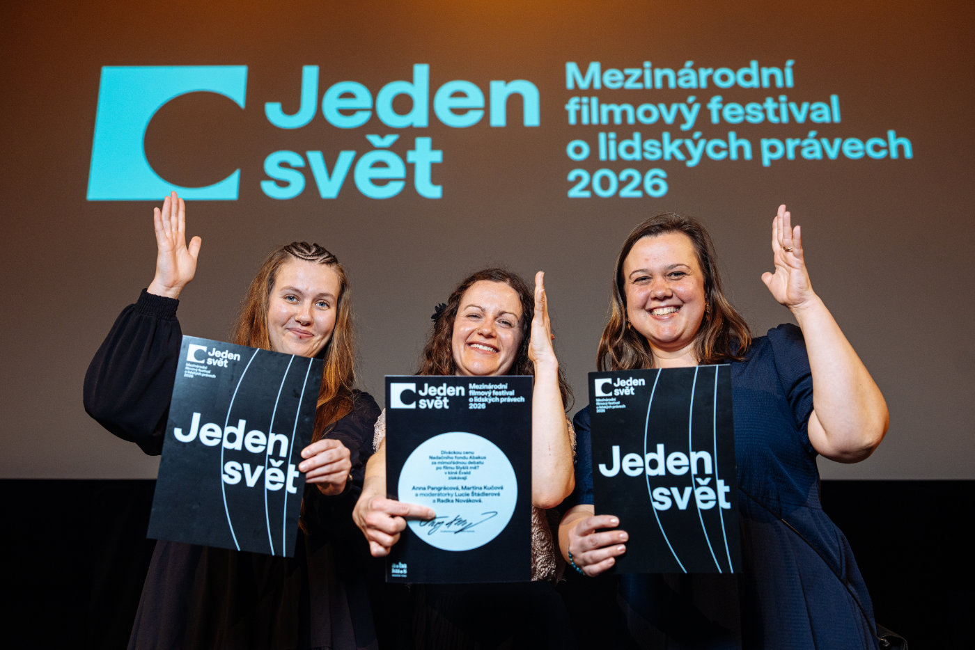 Tři ženy se usmívají a drží diplomy před plátnem s logem festivalu Jeden svět.