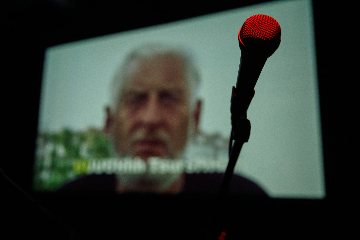 Červený mikrofon v popředí, v pozadí rozmazaný portrét muže na promítacím plátně.