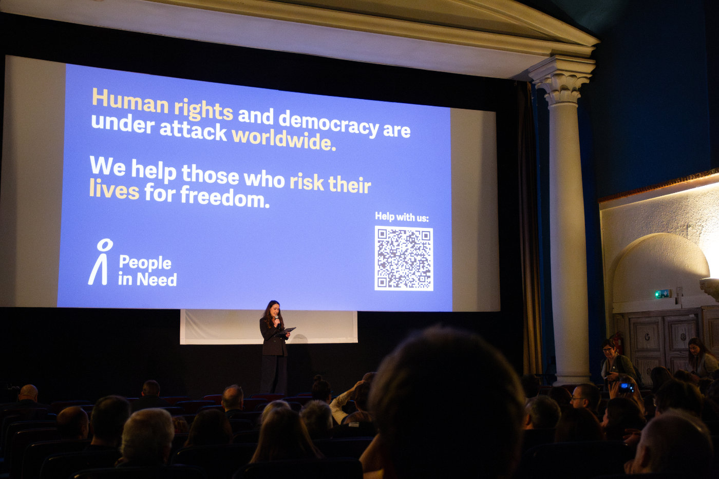 Žena stojí na pódiu před plátnem s prezentací o lidských právech a demokracii, v sále sedí diváci.