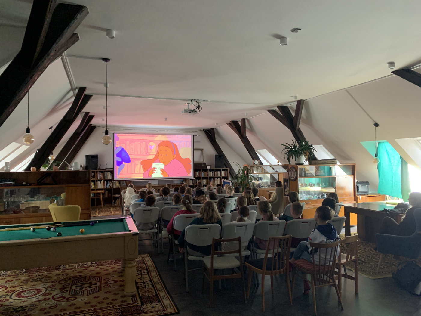 Lidé sedí v podkrovní místnosti a sledují promítání animovaného filmu na plátně.