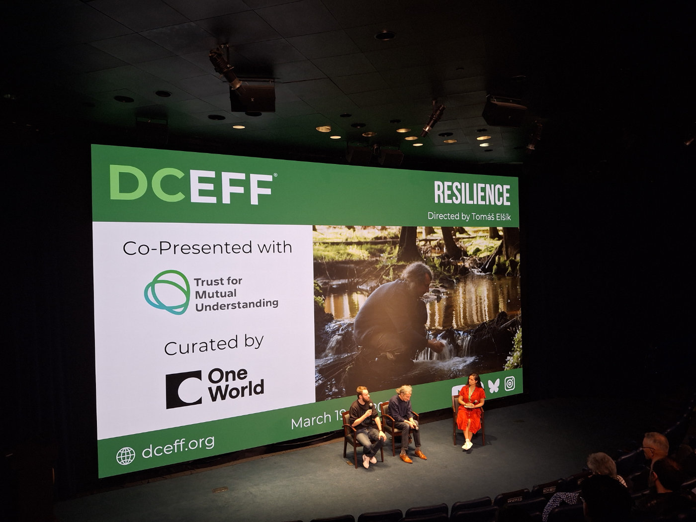 Tři lidé sedí na pódiu před velkým plátnem v kině, na kterém je promítána upoutávka na film „Resilience“ v rámci festivalu DCEFF.
